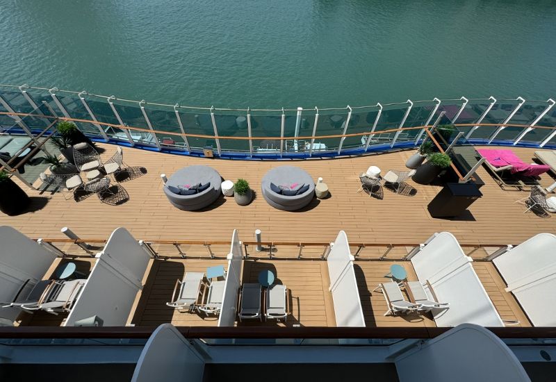 The image shows a view of the Cabana Deck on Princess Cruises' Sun Princess. The deck features private cabanas below, each equipped with lounge chairs and small tables, offering guests a semi-private outdoor space to relax. Above the cabanas, the deck extends with additional seating areas that include round daybeds, wicker chairs, and small tables arranged in a stylish and comfortable manner. The deck is surrounded by glass railings, providing unobstructed views of the ocean. The overall design emphasizes luxury and relaxation, making it an ideal spot for guests to enjoy the scenery and the sea breeze.