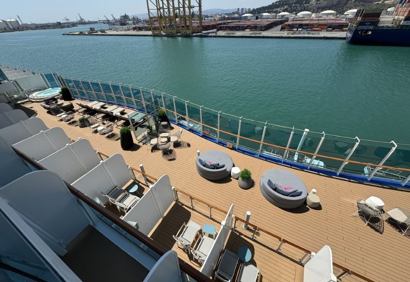 The image shows the Cabana Deck on Princess Cruises' Sun Princess. The deck features a series of private cabanas with lounge chairs and small tables, providing a semi-private outdoor space for guests. The deck is spacious with additional seating areas, including round daybeds and modern outdoor furniture, offering a comfortable and stylish environment for relaxation. The area overlooks the ocean, with a glass railing enhancing the view. The setting is ideal for sunbathing and enjoying the surrounding scenery while the ship is docked at a port.