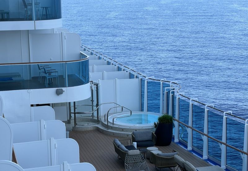 The image shows a section of the Cabana Deck on Princess Cruises' Sun Princess. The deck features private cabanas with white partition walls, providing an intimate outdoor space for guests. In the foreground, there is a hot tub surrounded by lounge chairs and modern outdoor seating, offering a comfortable and luxurious area to relax while enjoying the ocean view. The glass railings allow for unobstructed views of the sea, enhancing the serene atmosphere of the deck. The design combines privacy with scenic ocean views, making it an ideal spot for relaxation.