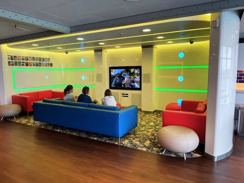 Children are engaged in video games at Camp Discovery on a Princess cruise ship, comfortably seated on colorful couches in a bright room with a large screen TV and modern, playful décor.