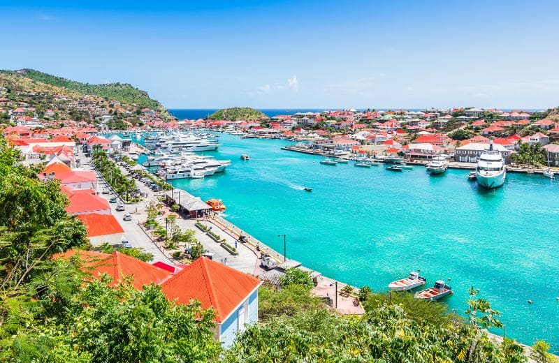 This image offers a stunning view of a harbor in St. Barts, a luxurious Caribbean destination known for its vivid blue waters and upscale atmosphere. The harbor is bustling with a variety of vessels, from small boats to larger yachts, all docked along a waterfront lined with charming red-roofed buildings. The landscape is lush with tropical vegetation, and the rolling hills in the background are dotted with colorful residential buildings, showcasing the island's vibrant architecture and scenic beauty. This setting perfectly encapsulates the luxurious and relaxed lifestyle that St. Barts is famous for.