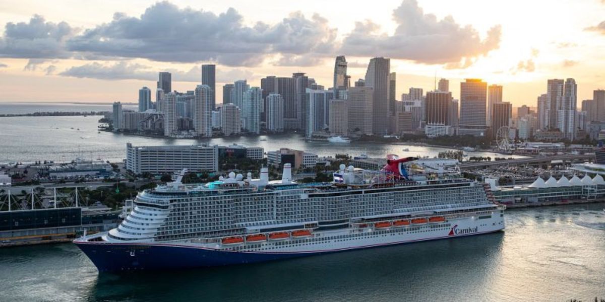 The image shows a Carnival cruise ship, "Carnival Celebration," docked in Miami with the city skyline in the background. The sun is setting, casting a warm glow over the high-rise buildings and the water, creating a scenic and vibrant atmosphere.