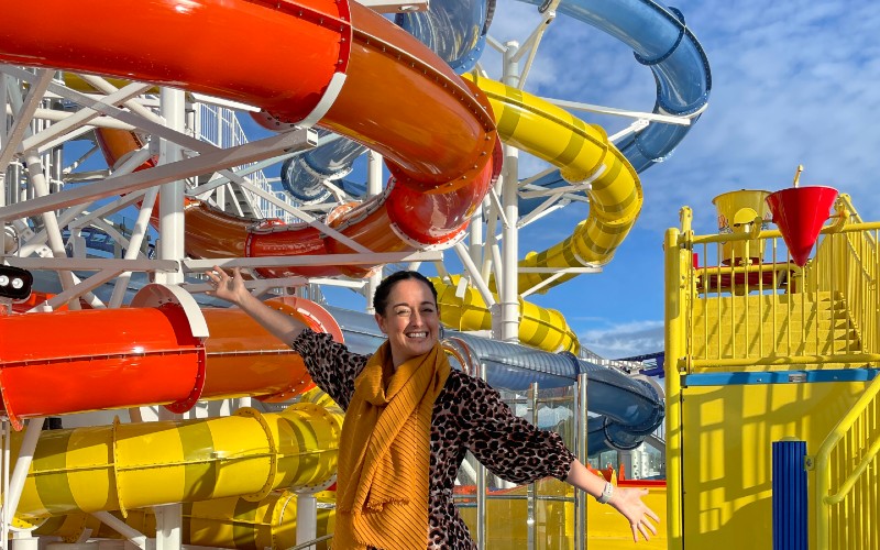Cruise Mummy in a leopard print coat and orange scarf smiles with arms outstretched in front of a colorful water slide structure at the Carnival Celebration water park. The water slides are vibrant in red, yellow, and blue, creating a lively backdrop under a clear blue sky.