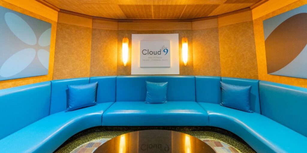 Inside the Cloud 9 Spa on a Carnival cruise ship, a tranquil lounge area with a sleek blue curved sofa is illuminated by soft wall sconces, creating a relaxing ambiance. The spa's logo is prominently displayed, inviting guests to unwind in this stylish and peaceful setting.