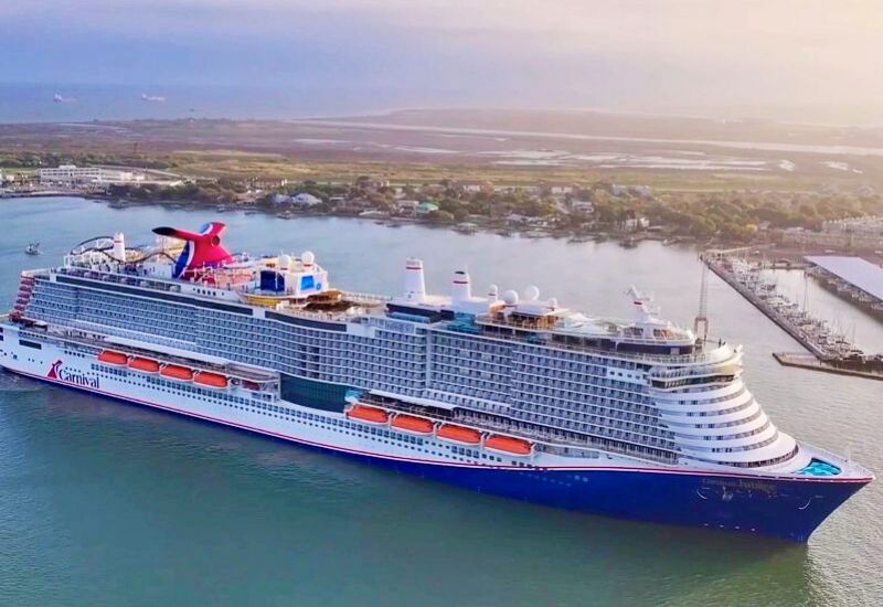 The Carnival Jubilee cruise ship majestically sails near Galveston, Texas, highlighted by the warm glow of sunset, with the island's waterfront and marinas creating a picturesque backdrop.