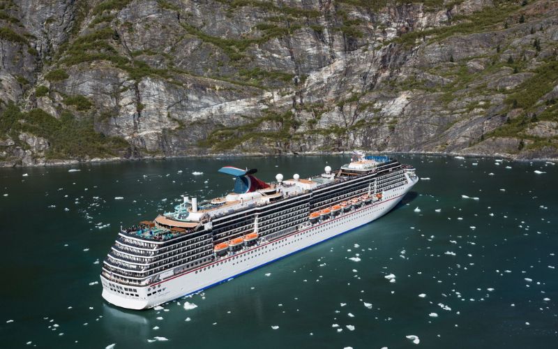 The Carnival Miracle cruise ship sails through icy waters, with a striking backdrop of sheer cliff faces. Small chunks of ice float nearby, adding to the sense of adventure in this secluded waterway.