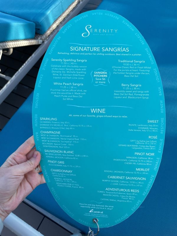 A hand holding the Serenity bar menu on a Carnival cruise ship, featuring a selection of signature sangrias and a wide range of wines, from sparkling to sweet, with detailed descriptions and prices, set against the backdrop of a serene turquoise lounger.