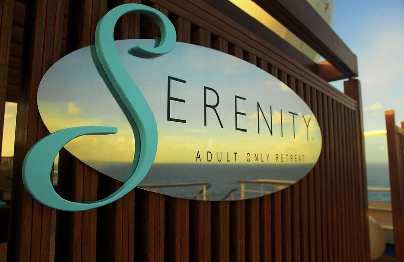 Close-up of the 'Serenity Adult Only Retreat' sign on a Carnival Cruise ship, featuring stylish turquoise script on a frosted glass panel set against a wooden background with a blurred ocean view at sunset in the distance.