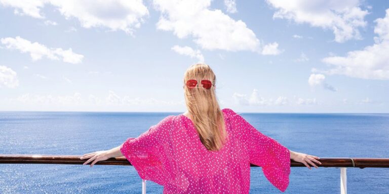 A woman in a bright pink polka dot dress stands at the railing of a Carnival Cruise ship, gazing out at the expansive ocean under a clear blue sky. Her long blonde hair is held back by red sunglasses, and the serene scene captures the tranquility of a solo cruise experience.