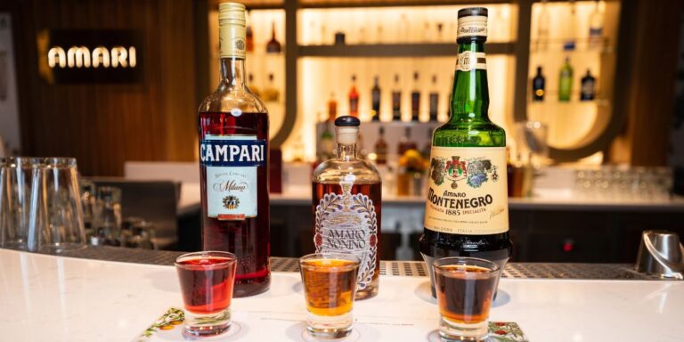 This image shows three bottles of liquor displayed on a bar counter aboard a Carnival Cruise ship. The bottles include Campari, Amaro Nonino, and Amaro Montenegro, each accompanied by a small glass of the corresponding drink in front of them. The background features a well-lit bar area with a variety of other bottles on shelves, and a sign that reads "AMARI." The setup highlights the selection of Italian liqueurs available on the cruise.