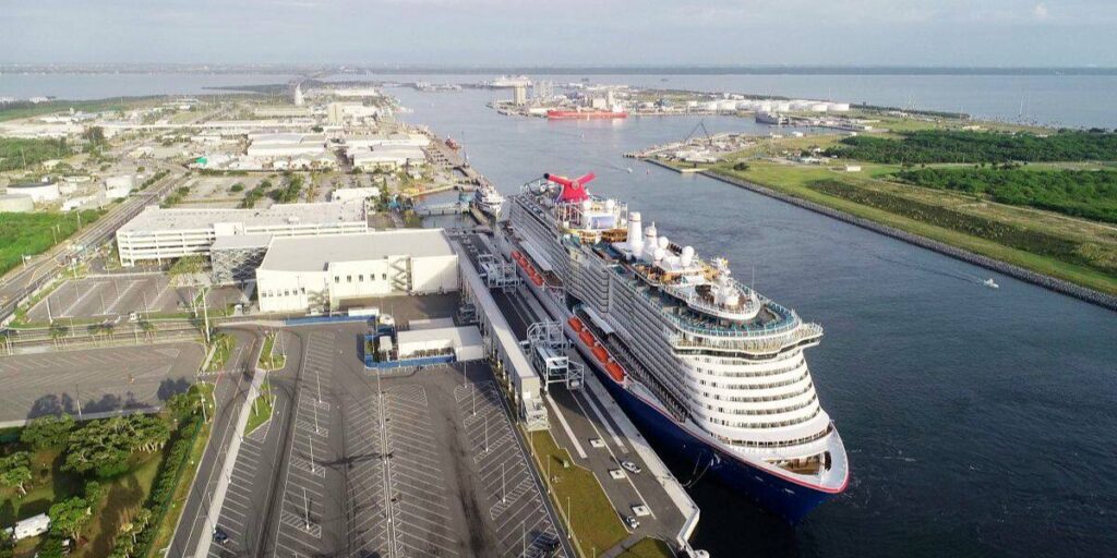 Aerial view of a Carnival cruise ship docked at a bustling port, with the ship's distinctive red funnel standing out against the sprawling industrial backdrop and calm blue waters.