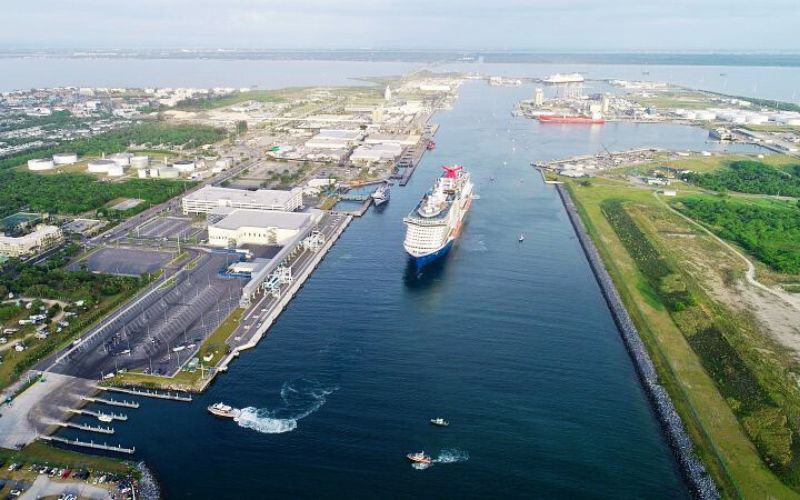 Aerial view of a Carnival cruise ship docked at Port Canaveral, Florida, with the vast expanse of the port infrastructure visible in the background, illustrating the scale and operation of a major cruise ship port.