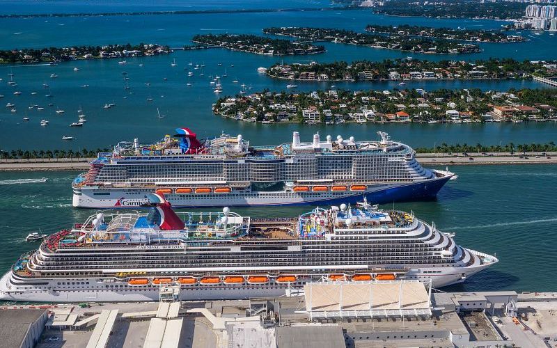 A stunning aerial shot of two Carnival cruise ships docked side by side in the Port of Miami, their blue and red funnels iconic against the picturesque backdrop of Miami’s coastal waterways and bustling marina life.