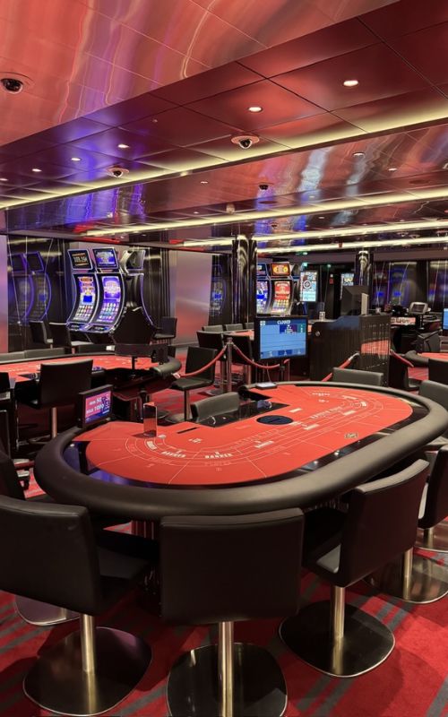 A vacant casino table awaits players in a cruise ship's casino, with the red baize of a baccarat table in the foreground and a row of slot machines in the background. The space is characterized by reflective red and purple metallic ceilings and patterned carpet, creating a vibrant gaming atmosphere.