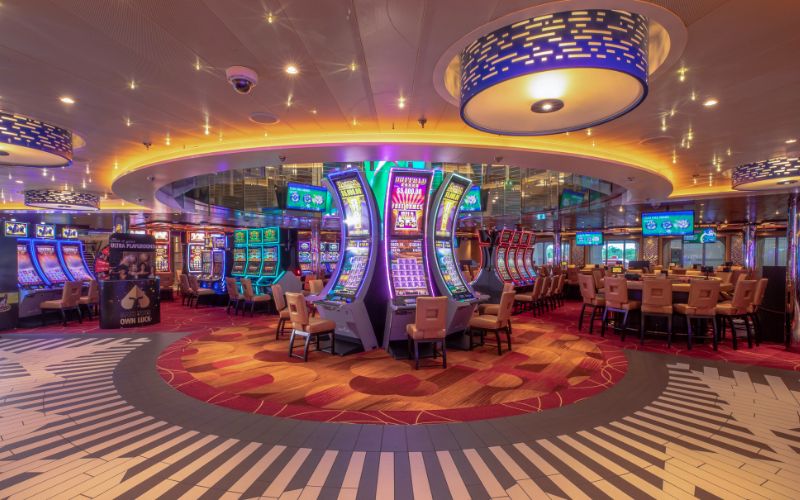 Vibrant casino floor on the Carnival Horizon, featuring an array of colorful slot machines and gaming tables, with dynamic lighting overhead and a bold, patterned carpet adding to the lively atmosphere.