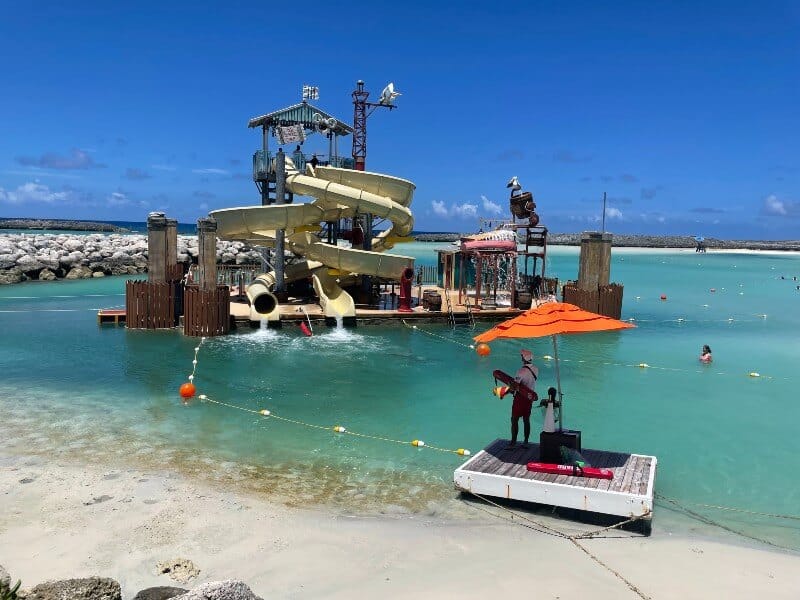 Visitors enjoy the sun-soaked waterslides at Castaway Cay, with a musician playing steel drums under an orange umbrella on the white sandy beach.