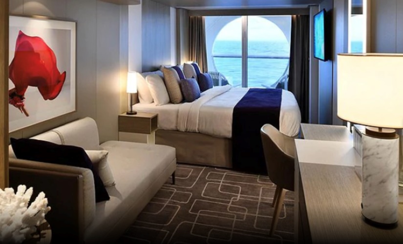 Elegant interior of a Deluxe Porthole Balcony Stateroom on a Celebrity Cruise ship, featuring a plush bed with ocean views, a comfortable sitting area, and modern decor with artistic wall art and geometric carpet design.