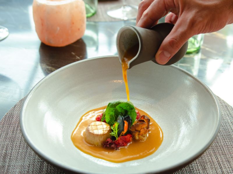 A chef artfully presents a gourmet dish at Eden Restaurant on a Celebrity Cruise, pouring a vibrant orange sauce over a beautifully plated scallop with accompanying garnishes, symbolizing the fine dining experience at sea.