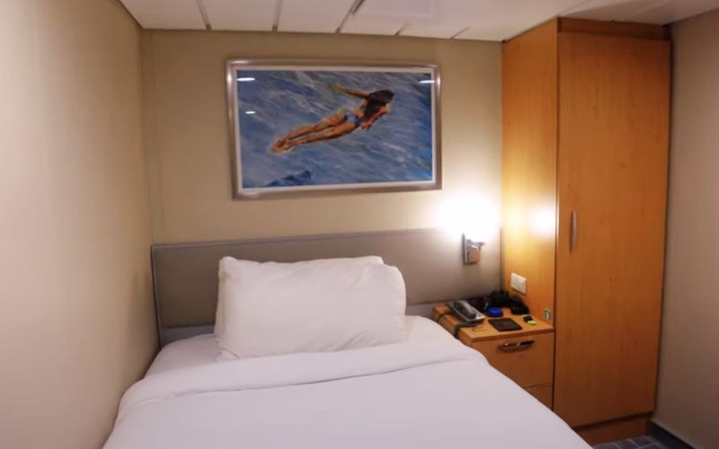 A solo cabin on the Celebrity Silhouette, designed for single travelers, featuring a comfortable bed, warm lighting, and a framed picture of a swimmer, creating a cozy and inviting space for relaxation at sea.