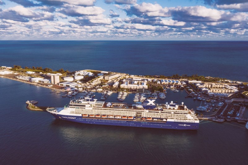 The Celebrity Summit cruise ship docked at a port, captured from an aerial perspective, showcasing the elegant vessel with its distinctive 'X' funnel next to the marina with clear blue waters extending into the horizon.