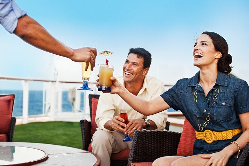 Guests aboard a Celebrity Cruises ship enjoying a toast with refreshing drinks, featuring a man and a woman laughing and smiling while they receive their beverages, with the calm ocean and clear skies in the background.