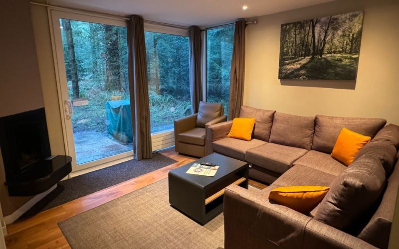 Cozy woodland-themed living room with a large corner sofa adorned with orange cushions, a simple black coffee table, and a forest view through floor-to-ceiling windows. A wall-mounted fireplace and a framed picture of a forest add to the tranquil, nature-inspired ambiance.