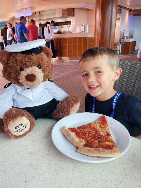 A smiling young boy sitting at Princess Cruises' World Fresh Marketplace next to a teddy bear dressed in a ship captain's uniform, with a slice of pizza on a plate in front of them.