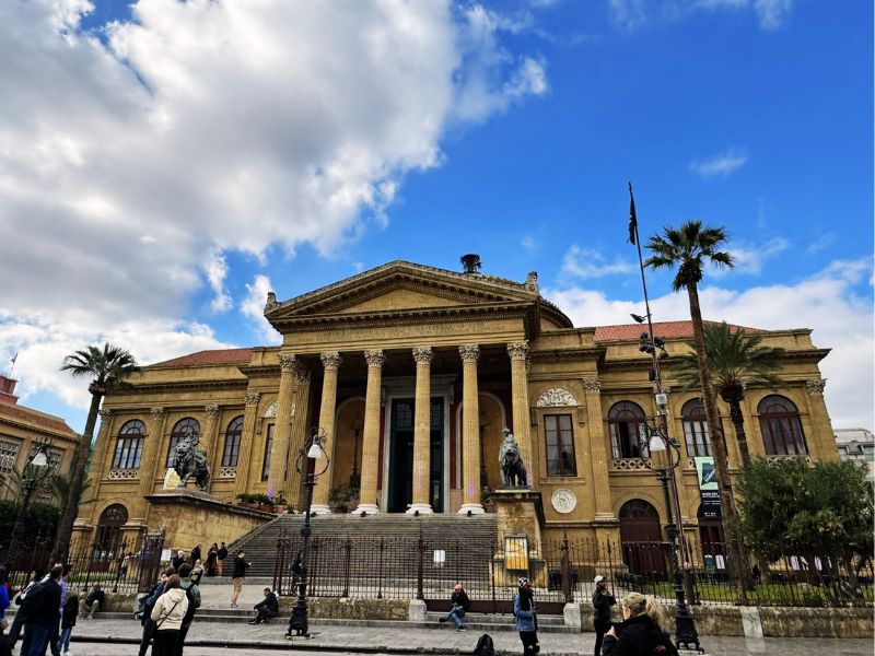 The majestic Teatro Massimo in Palermo, Sicily, stands under a bright blue sky with fluffy clouds, with its neoclassical architecture, grand entrance steps, and statues, as people walk and sit in the square in front.