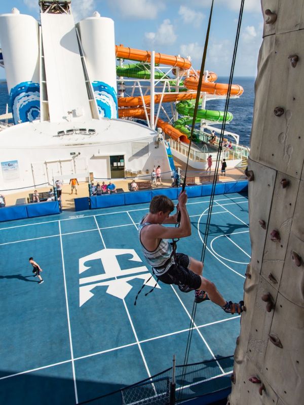 An adventurous guest scales the climbing wall on Royal Caribbean's Liberty of the Seas, poised against a backdrop of exhilarating water slides and the expansive ocean. Below, passengers enjoy a game of basketball on the ship's sports court, offering a snapshot of the diverse and active entertainment options available onboard.