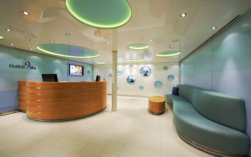 The reception area of Cloud 9 Spa on a Carnival cruise ship exudes a modern and calming vibe with a curved wooden reception desk, sleek blue curved sofa, and ambient lighting from circular ceiling fixtures. The spa's theme of relaxation is echoed in the circular wall mirrors and soft color palette.