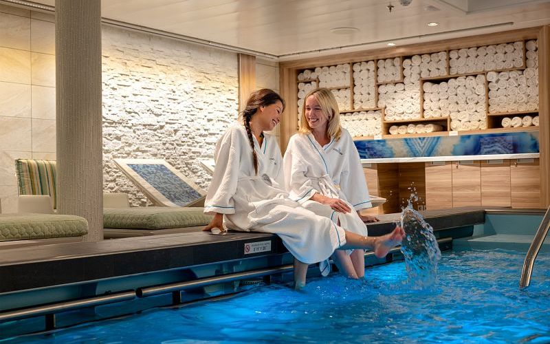 Two women enjoy a moment of relaxation at the Cloud 9 Spa on a Carnival cruise ship, sitting by a therapeutic pool. They are clad in plush white robes, laughing together in a luxuriously designed spa interior with a backdrop of textured white walls and neatly stacked towels.