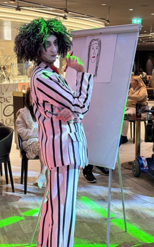 A performer in a black and white striped suit with a curly green wig poses beside an easel with a simple line drawing of a face, creating a whimsical and artistic atmosphere in MSC Euribia cruise ship's bustling indoor promenade