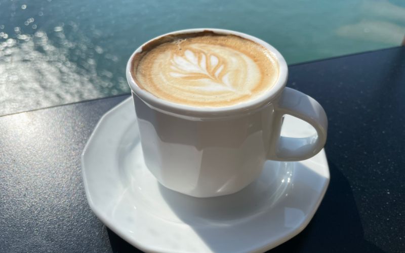 A cup of cappuccino with latte art on top, presented on a saucer, with the ocean glistening in the background, capturing a serene coffee moment by the sea.