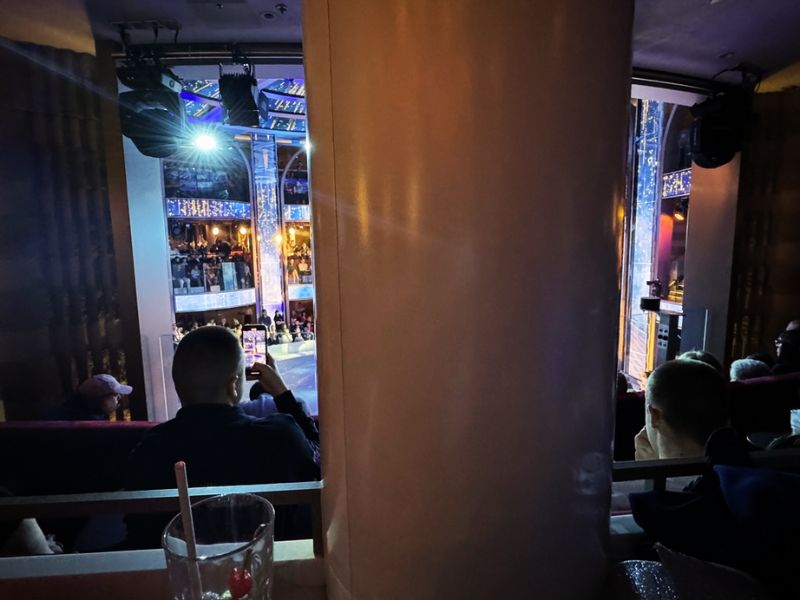 A view from a secluded spot inside the Colosseum on Costa Smeralda, capturing the audience and stage from an upper level, with the spotlight shining and a person in the foreground taking a photo of the performance.