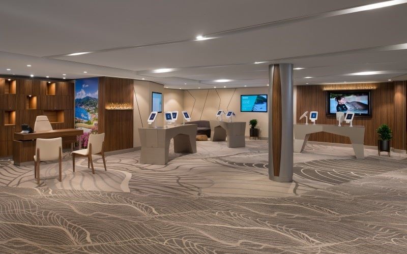 The Concierge Desk and Shore Excursion area on Celebrity Edge, designed with a contemporary aesthetic, featuring interactive screens, elegant furniture, and a calming neutral color palette complemented by the soft, wave-patterned carpet.