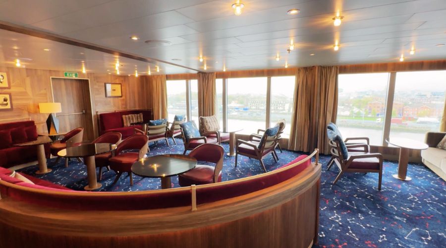 Constellation Lounge aboard the Seabourn Venture, a spacious and elegantly appointed area designed for relaxation and socializing. The lounge is furnished with plush seating options including velvet sofas and leather armchairs, arranged around sleek coffee tables. Rich wooden paneling adds warmth to the space, while the large windows offer abundant natural light and panoramic views of the port outside. This lounge provides an inviting atmosphere where guests can gather to enjoy drinks, conversation, and the beautiful scenery as they cruise. The decor, highlighted by a tasteful marine-themed carpet, enhances the nautical vibe of the ship.