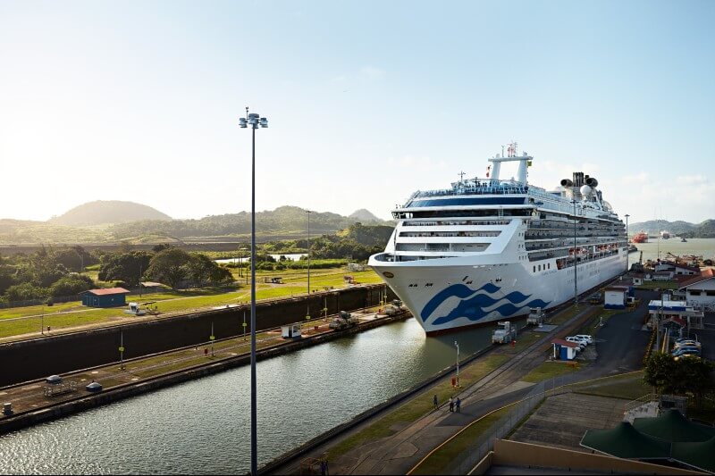 This image showcases the Coral Princess cruise ship navigating through the Panama Canal. The ship is surrounded by lush greenery, rolling hills, and the iconic canal locks, highlighting the engineering marvel of this passageway. The scene captures the grandeur of the cruise experience against a backdrop of natural beauty and impressive infrastructure.