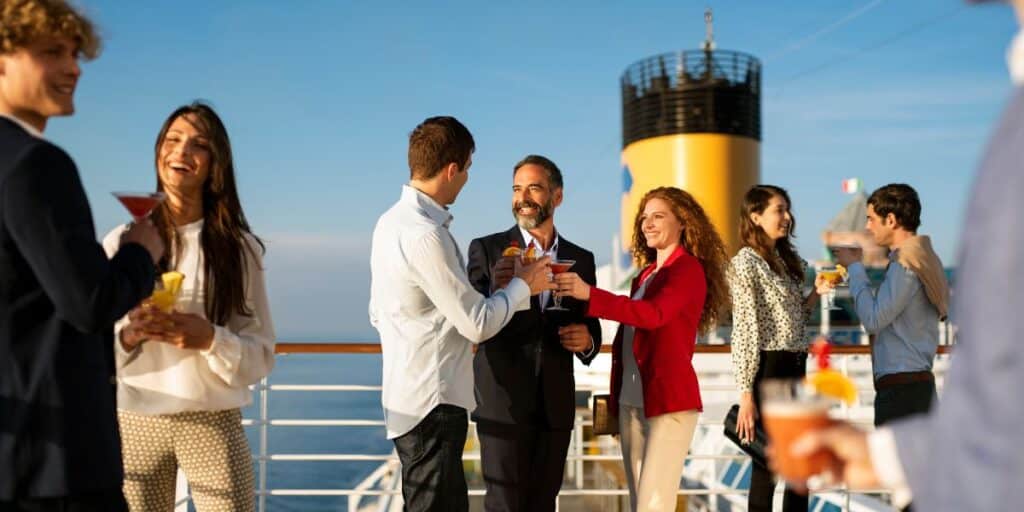 A group of people enjoying a social event on the deck of a cruise ship, with a clear blue sky above and the ship's yellow funnel in the background. They are dressed in smart casual attire, holding cocktails, and engaging in cheerful conversation.