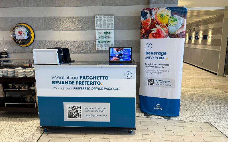 Beverage info point in a cruise ship setting, featuring a blue stand with text 'Scegli il tuo PACCHETTO BEVANDE PREFERITO. Choose your PREFERRED DRINKS PACKAGE.' and a QR code, accompanied by a vibrant banner of refreshing drinks, promoting onboard amenities.