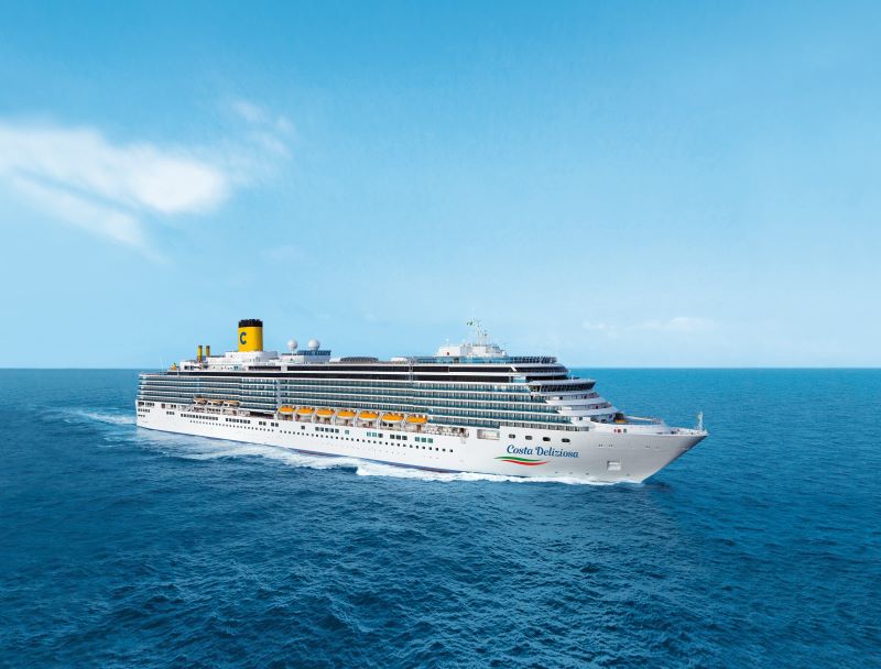 The Costa Deliziosa cruise ship captured from a side angle as it cruises on the deep blue ocean. Its white structure adorned with rows of balconies and distinctive yellow lifeboats is complemented by the ship's yellow funnel featuring a blue 'C' emblem, cruising under a bright blue sky with light wispy clouds.
