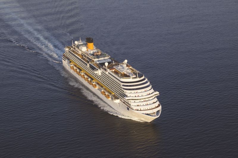 An aerial view of the Costa Diadema cruise ship traveling on a sunlit sea, its wake forming a V-shaped pattern in the water. The ship's deck is lined with yellow lifeboats, and its large yellow funnel stands out with the letter 'C' against the deep blue of the ocean.