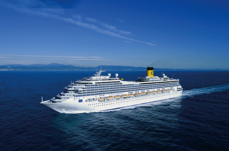 The Costa Fortuna cruise ship is showcased in full sail across the open sea, captured from a high angle showcasing its white structure, multiple decks with rows of windows, and a prominent yellow funnel with the 'C' logo. In the background, a distant coastline with mountains can be seen under a clear blue sky.