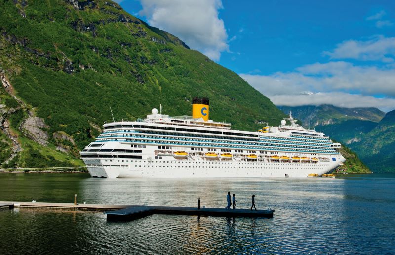 The Costa Fortuna cruise ship moored in a fjord, surrounded by steep green mountains. The ship's white exterior with yellow lifeboats and prominent yellow funnel with a 'C' logo stands out in the natural beauty of the setting, while a small pier with people adds scale to the serene scene.