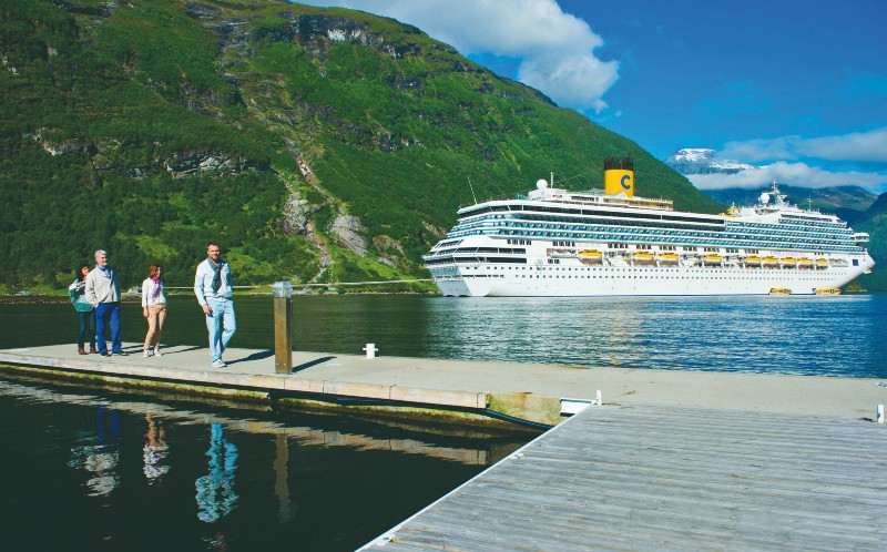 A group of tourists walking on a wooden pier in Norway, with the Costa Fortuna cruise ship docked in the background against a backdrop of lush green mountains under a blue sky