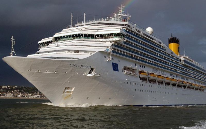 The Costa Magica cruise ship embarks on a voyage, its white and yellow colors standing out against a dramatic sky with a faint rainbow visible in the distance.