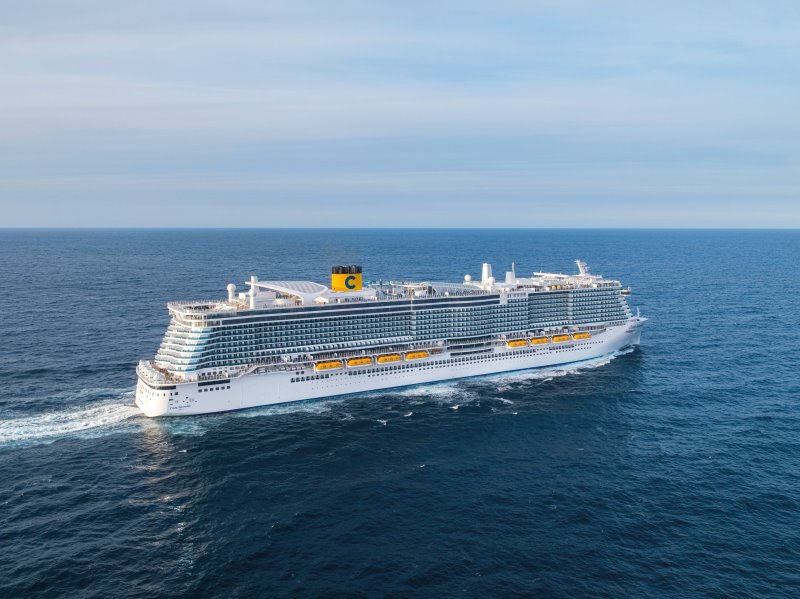 Aerial view of the Costa Smeralda cruise ship sailing on the open sea, featuring multiple decks with yellow lifeboats, a distinctive yellow funnel with a blue 'C' logo, and a wake of white foam on the blue water.