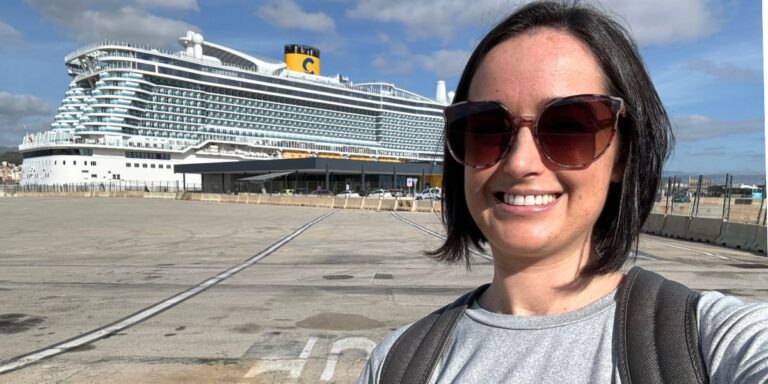 Cruise Mummy with sunglasses smiling at the camera with the Costa Smeralda cruise ship docked in the background under a partly cloudy sky.