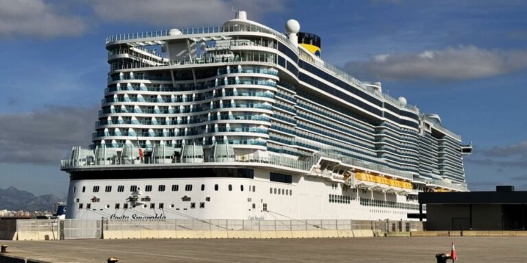 The Costa Smeralda cruise ship docked in a port on a clear day, its white and blue balconies visible against the backdrop of a blue sky with fluffy clouds and distant mountains.