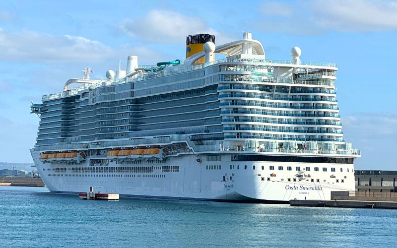 The Costa Smeralda cruise ship docked at a port, showcasing its white multi-deck structure with distinctive yellow and blue funnel, set against a backdrop of calm blue water and a partly cloudy sky.