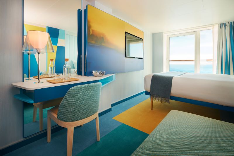 A bright and modern cabin on the Costa Toscana cruise ship, featuring vibrant blue and yellow decor. The room includes a comfortable bed, a desk with a stylish chair and lamp, and a wall-mounted TV. Large windows offer a beautiful ocean view, adding to the room's cheerful and inviting ambiance.